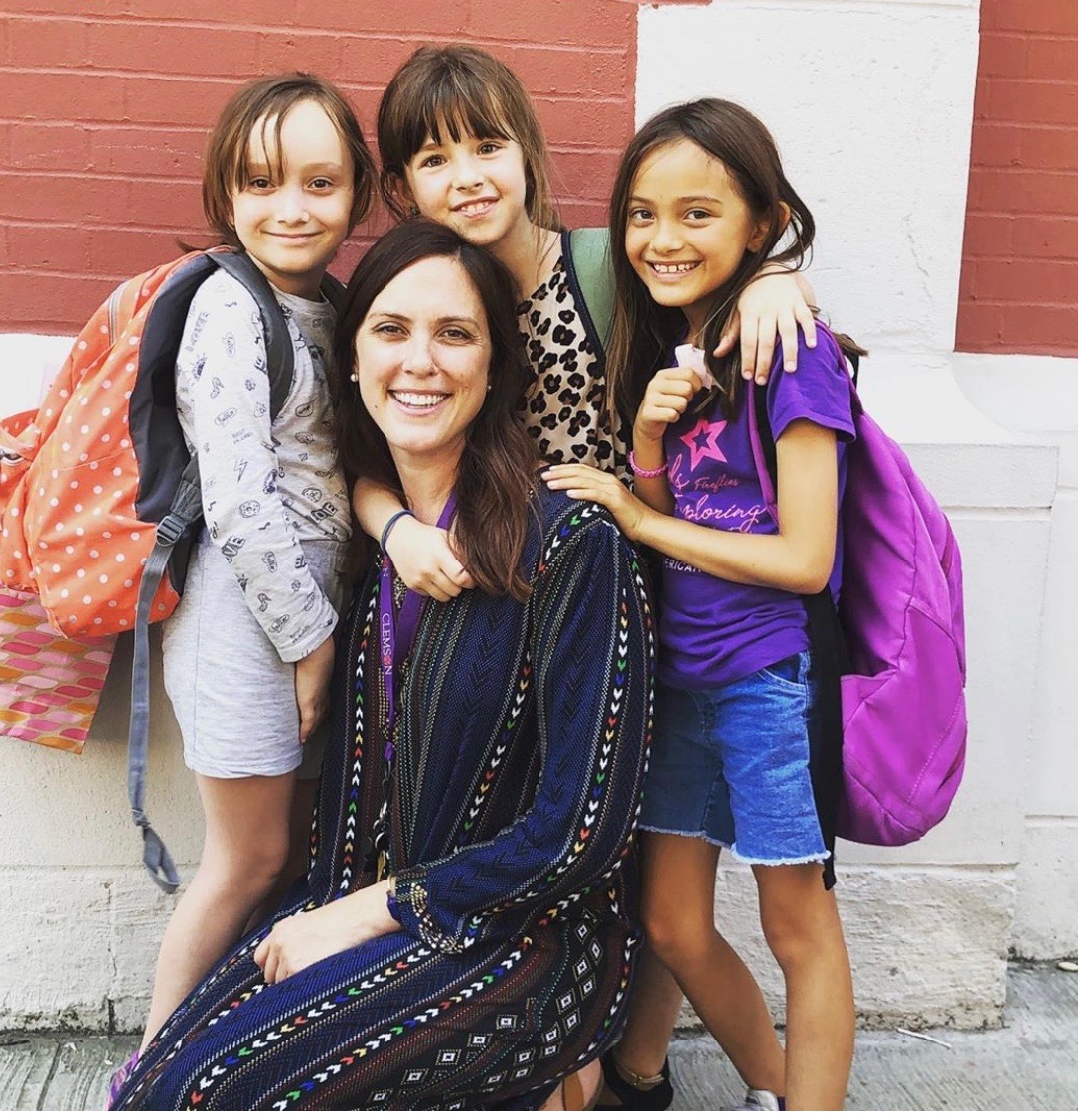 A woman kneeling in front of three young girls with backpacks, all smiling for a photo in front of a brick wall.